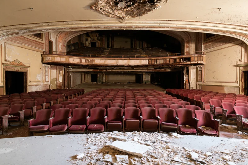Rows of red velvet seats in a decaying theater interior with crumbling walls and debris on the floor