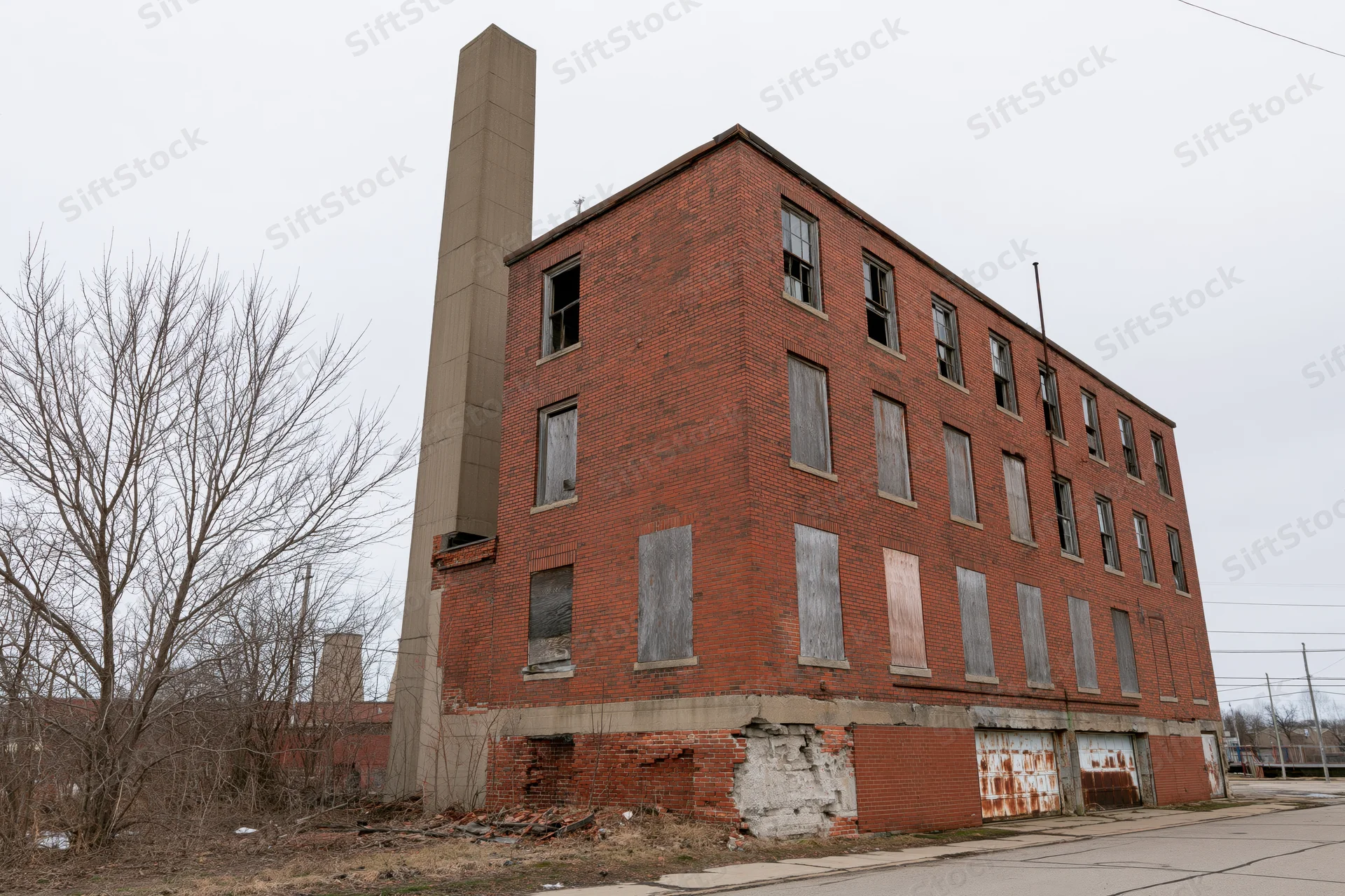 Abandoned three-story red brick industrial factory building with boarded windows and tall chimney on overcast day
