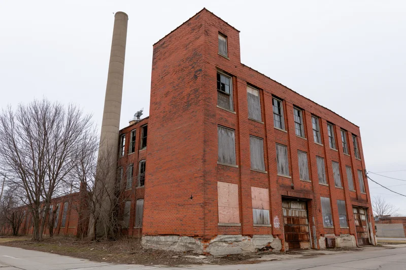 Abandoned three-story red brick industrial factory building with boarded windows and tall chimney on overcast day