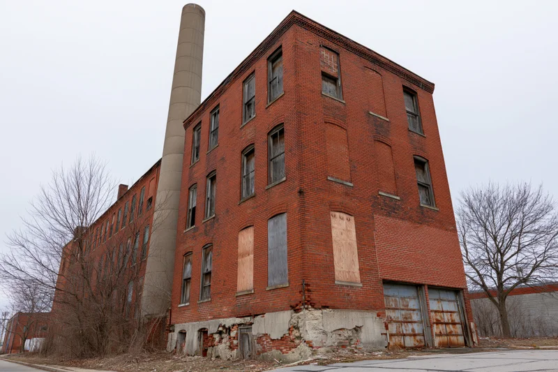 Abandoned three-story red brick industrial factory building with boarded windows and tall chimney on a gray winter day