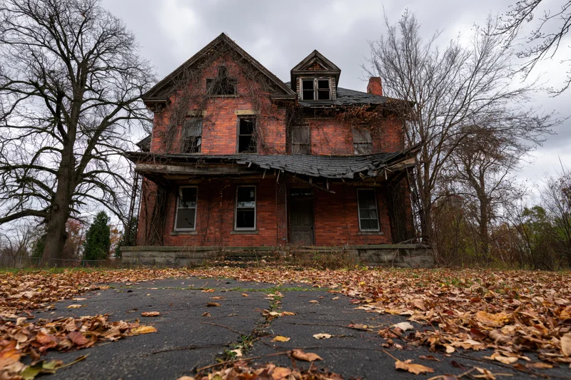 Abandoned two-story brick house with broken windows and dead vines overgrown in autumn landscape
