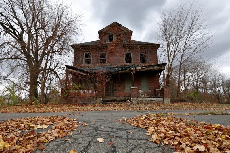 Abandoned two-story brick house overgrown with dead vines and surrounded by fallen autumn leaves on a cracked pavement under a cloudy sky