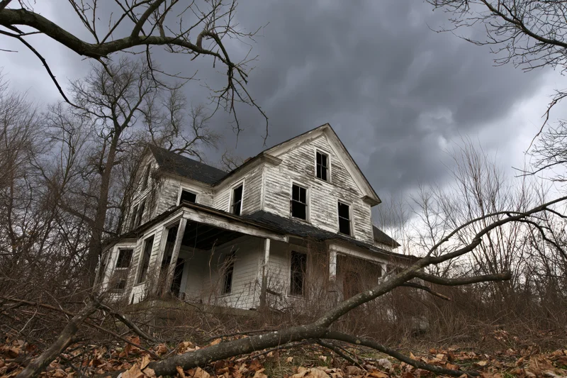 Abandoned two-story clapboard farmhouse with collapsed front porch surrounded by bare trees and fallen leaves under stormy sky