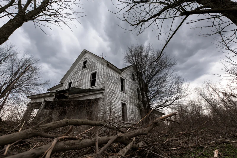 Abandoned two-story clapboard farmhouse with collapsed front porch surrounded by bare trees and overgrown brush under a cloudy sky