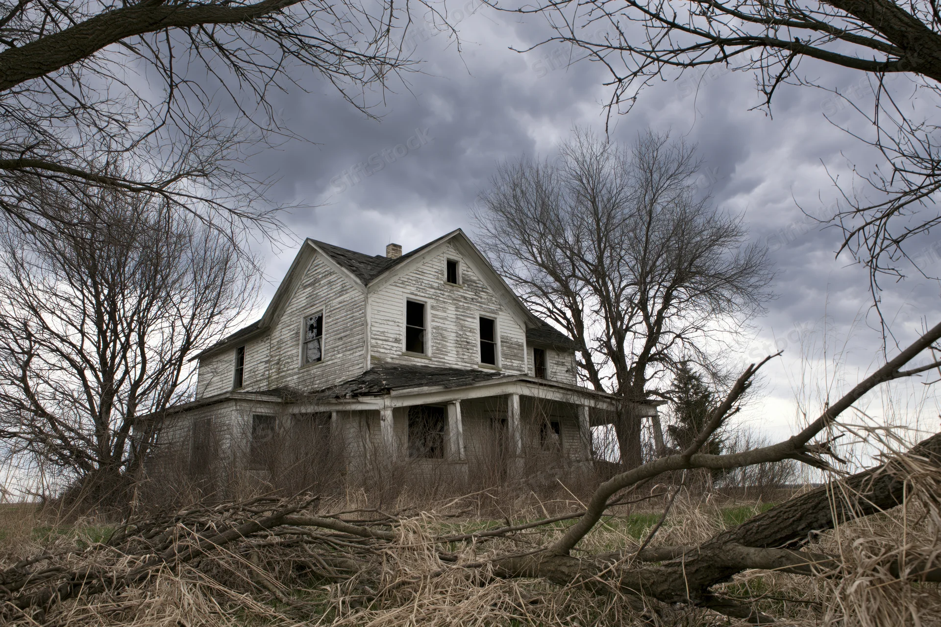 Abandoned two-story clapboard farmhouse with collapsed front porch surrounded by bare trees and overgrown field under cloudy sky