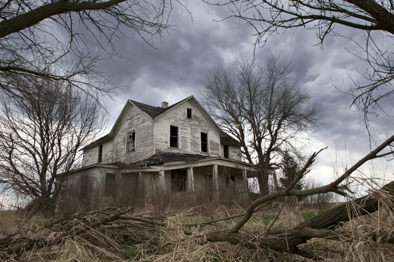 Abandoned two-story clapboard farmhouse with collapsed front porch surrounded by bare trees and overgrown field under cloudy sky