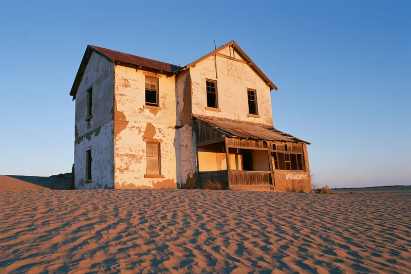 Abandoned two-story colonial house with crumbling plaster walls standing in desert sand under clear blue sky
