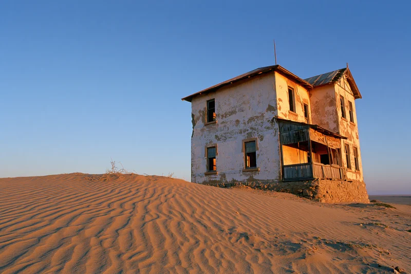 Abandoned two-story colonial house with crumbling plaster walls on a sand dune under clear blue sky at sunset