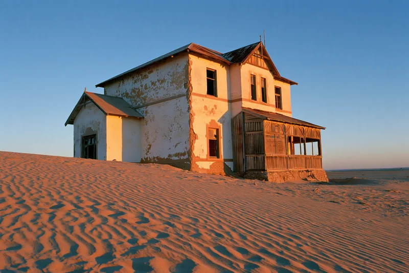 Abandoned colonial house with crumbling plaster walls and wooden porch surrounded by sand dunes under clear blue sky