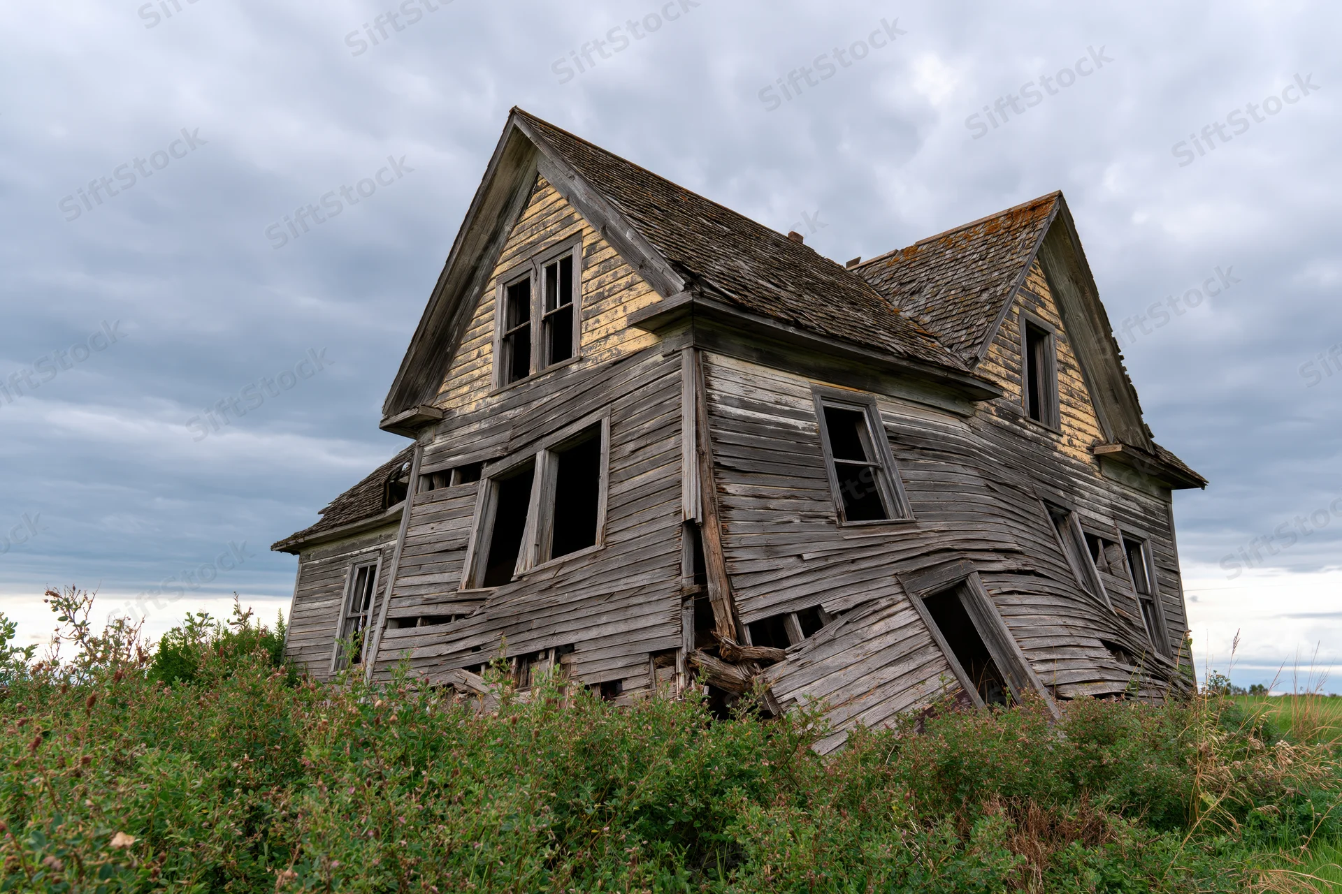 Abandoned two-story wooden farmhouse collapsing under weathered decay in overgrown rural field