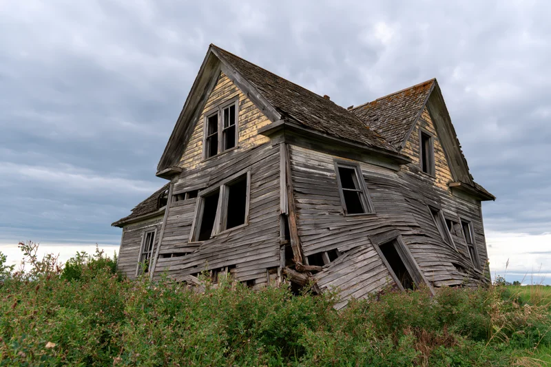 Abandoned two-story wooden farmhouse collapsing under weathered decay in overgrown rural field