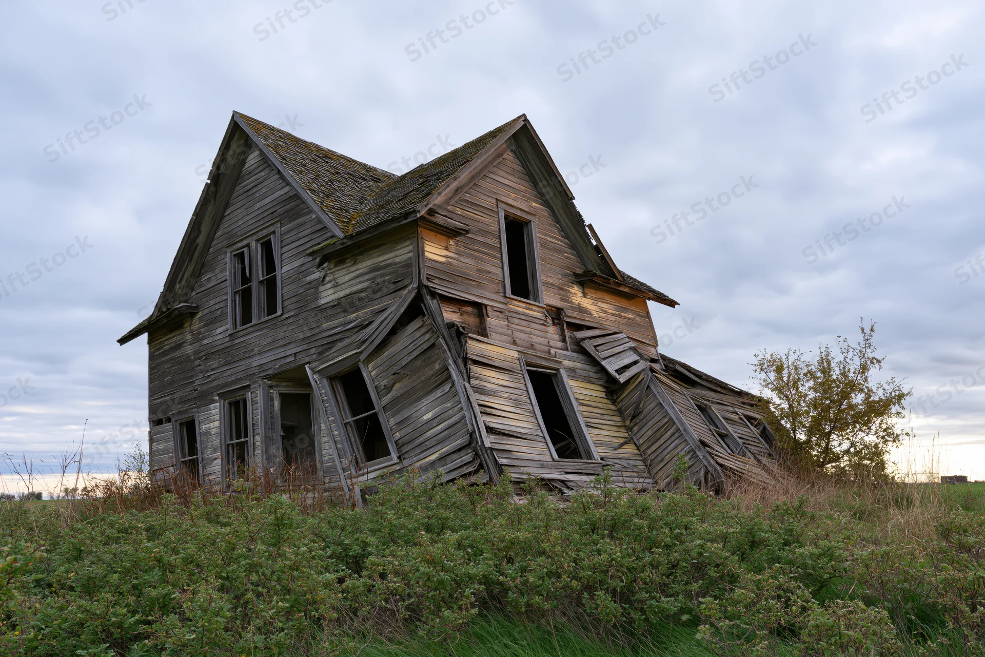 Collapsing two-story farmhouse in overgrown field under cloudy sky