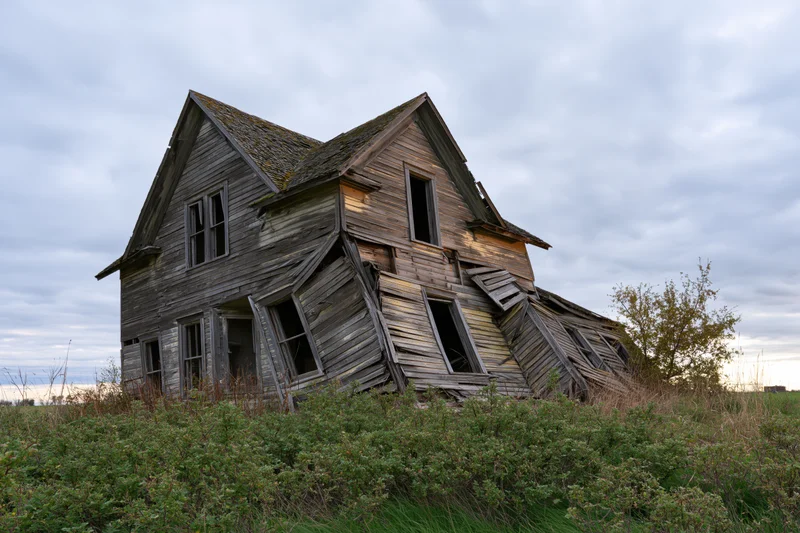 Collapsing two-story farmhouse in overgrown field under cloudy sky
