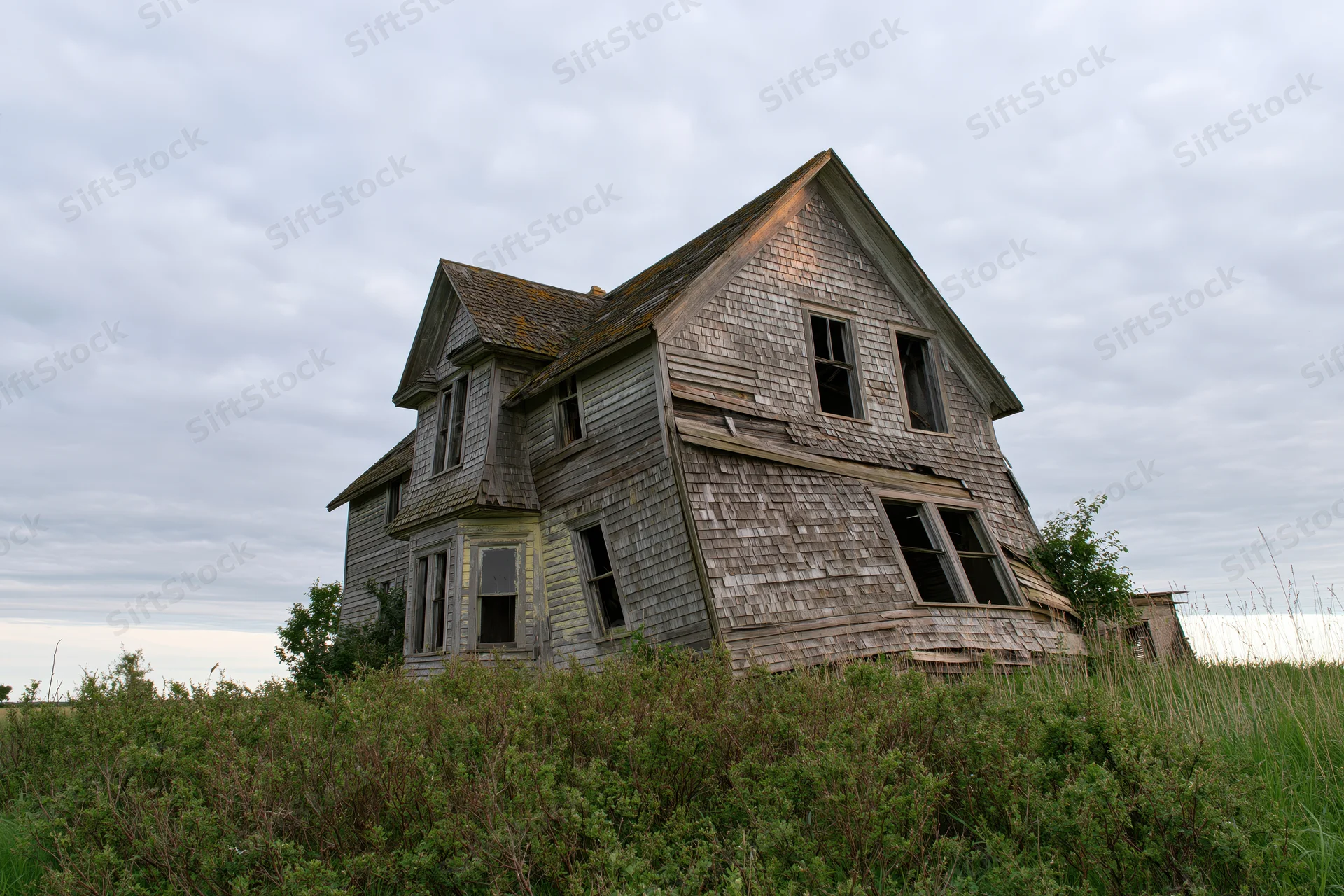 Abandoned two-story farmhouse collapsing in overgrown field under cloudy sky