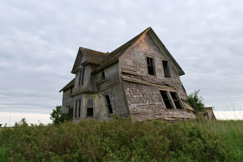 Abandoned two-story farmhouse collapsing in overgrown field under cloudy sky