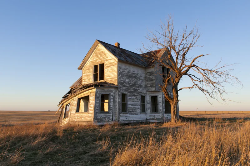 Abandoned two-story farmhouse with collapsed porch roof in a grassy field at sunset