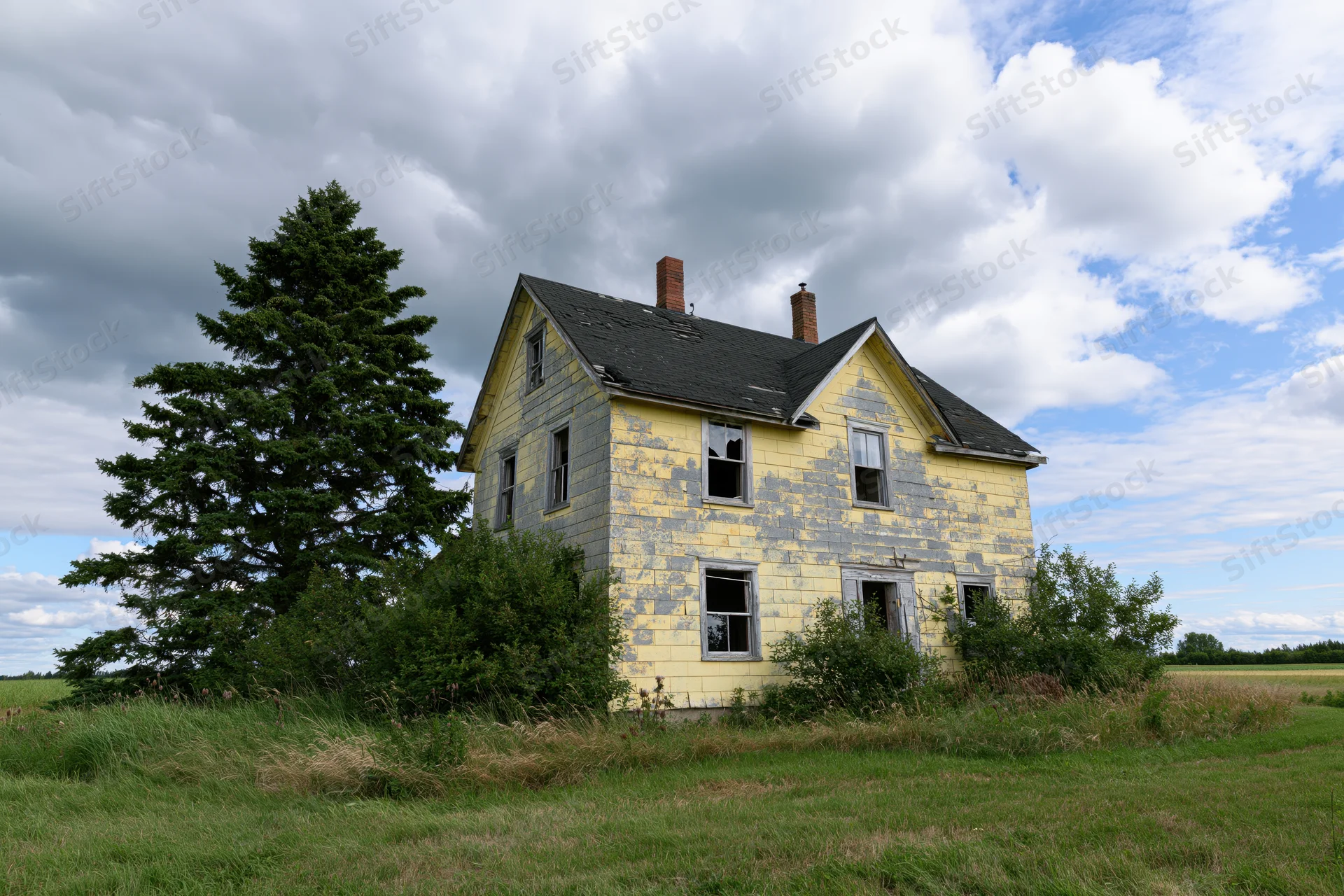 Abandoned two-story farmhouse with peeling yellow paint and broken windows in a grassy field under cloudy sky