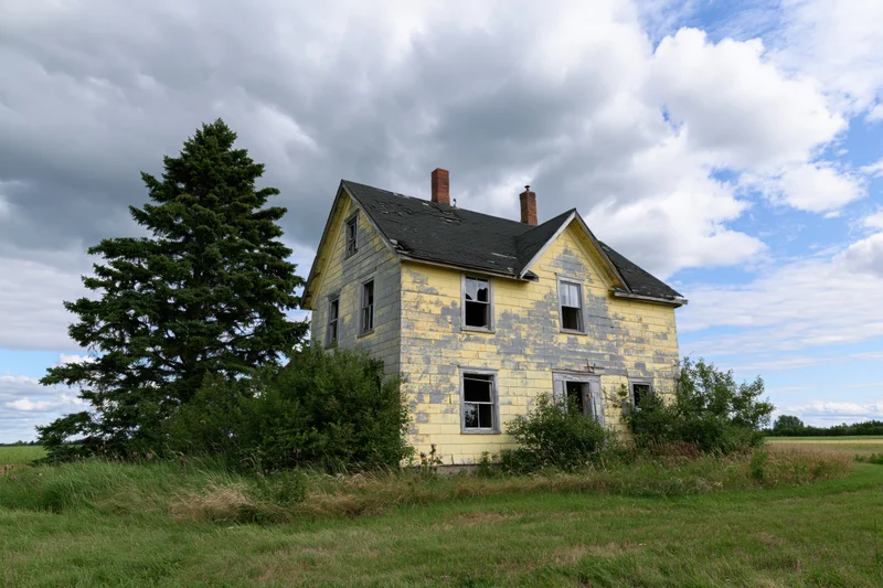 Abandoned two-story farmhouse with peeling yellow paint and broken windows in a grassy field under cloudy sky