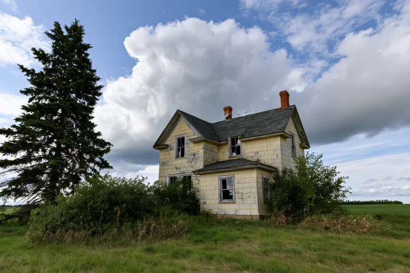 Abandoned two-story farmhouse with faded yellow shingle roof and broken windows surrounded by overgrown grass and trees in a rural field under a cloudy blue sky