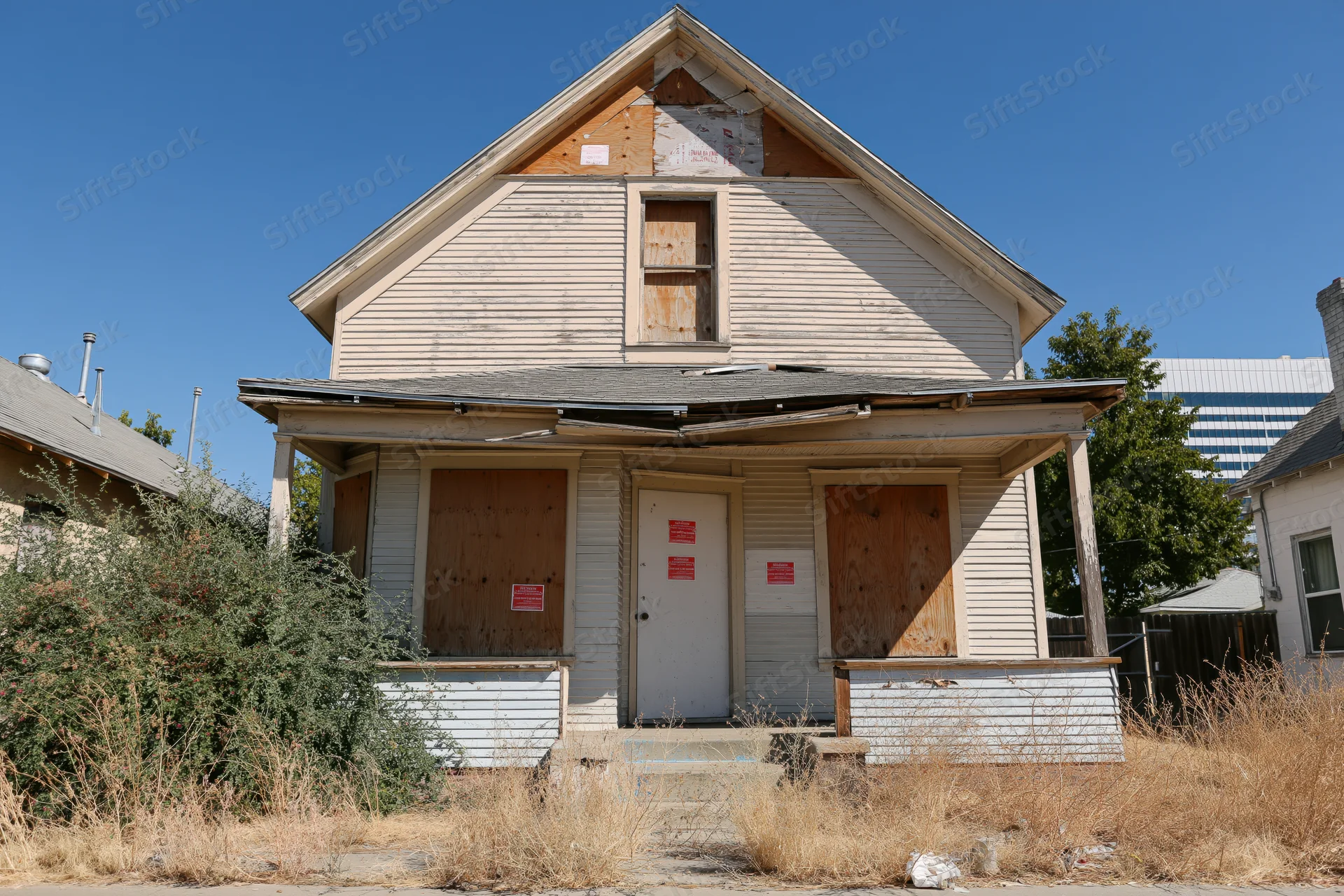 Abandoned two-story house with boarded windows and overgrown yard under clear blue sky