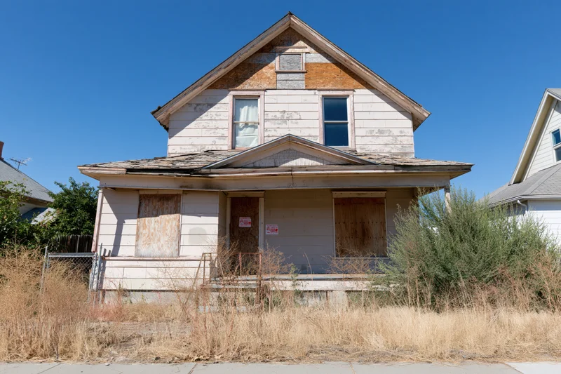 Abandoned two-story house with boarded windows and overgrown yard under clear blue sky