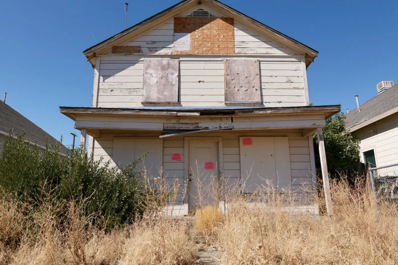 Abandoned two-story house with boarded windows and overgrown yard under clear blue sky