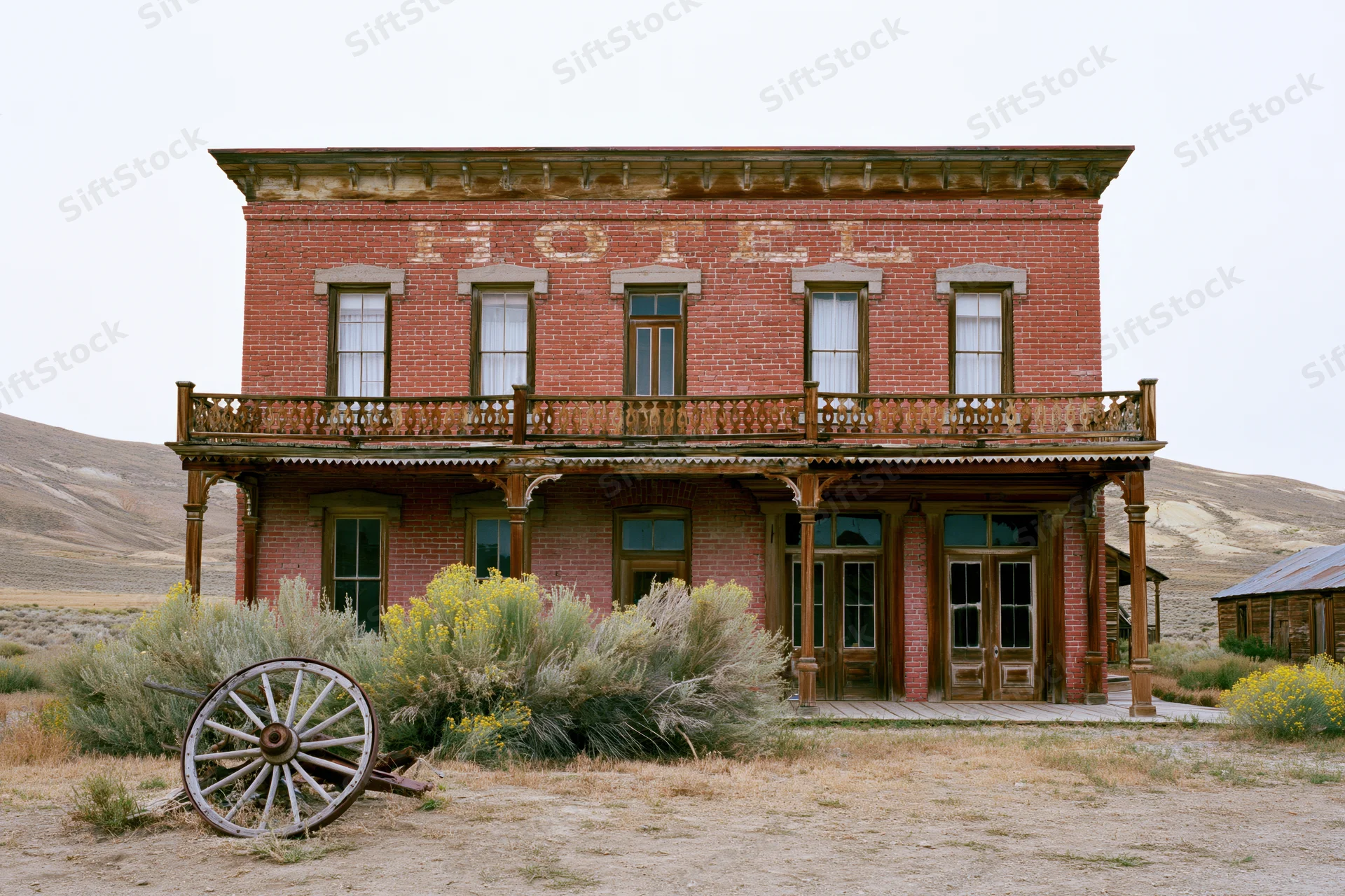 Abandoned two-story red brick hotel with wooden balcony and wagon wheel in front, set in a dry desert landscape