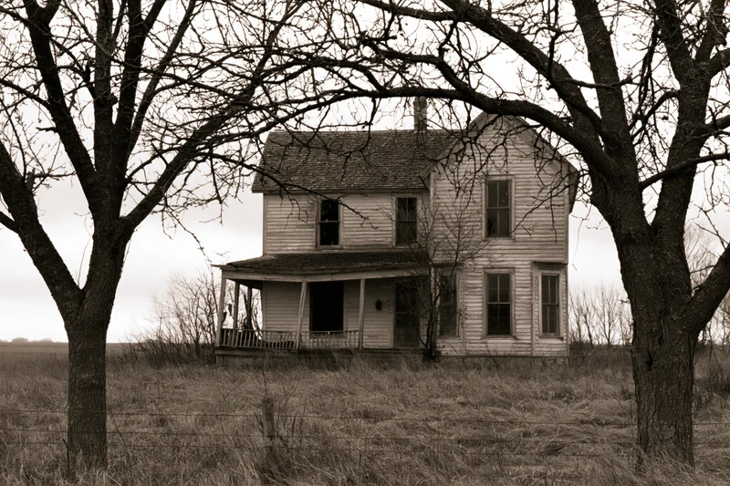 Abandoned two-story wooden farmhouse with sagging porch in a rural field framed by bare trees