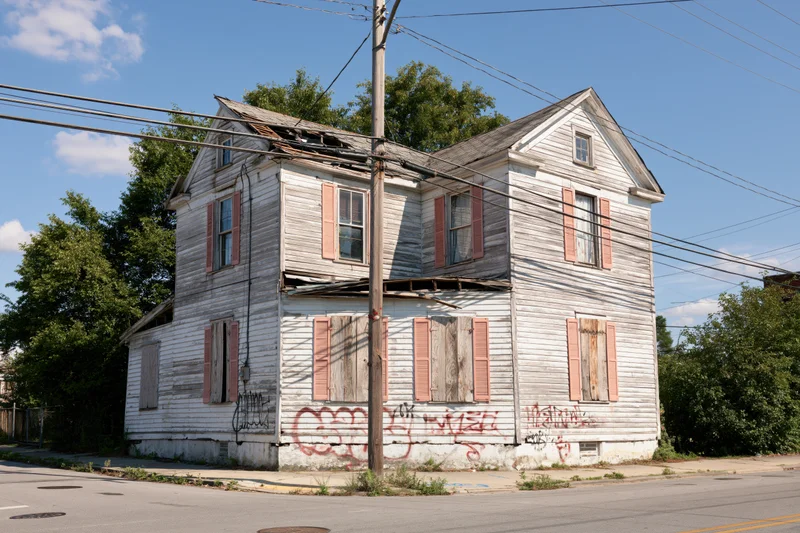Weathered two-story wooden house on a street corner with boarded windows and graffiti