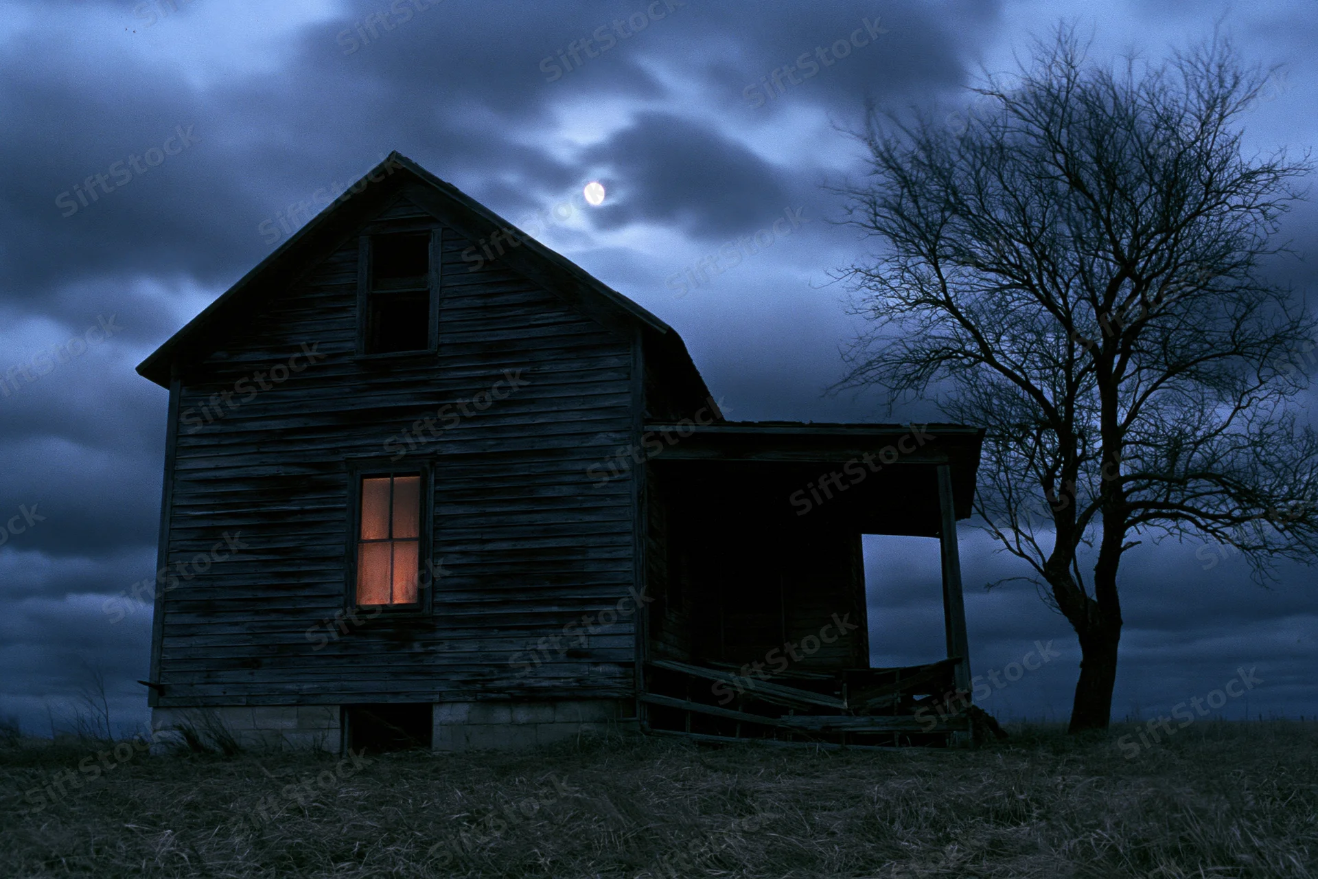 Abandoned wooden farmhouse with lit window at night in open field under cloudy sky with moon and bare tree