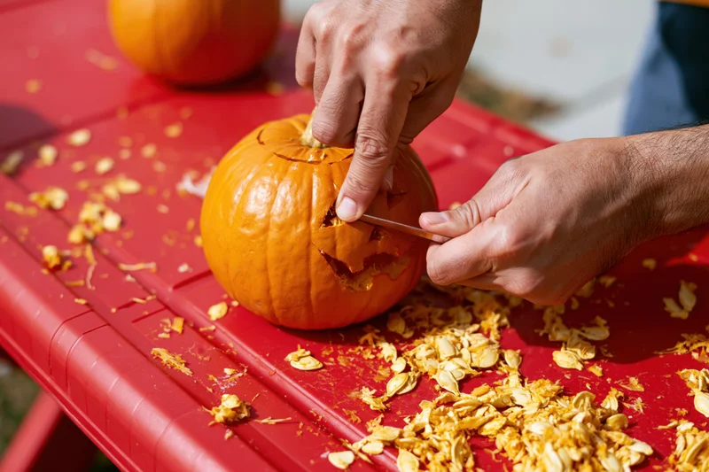 Hands carving a jack-o-lantern face into a small orange pumpkin on a red table