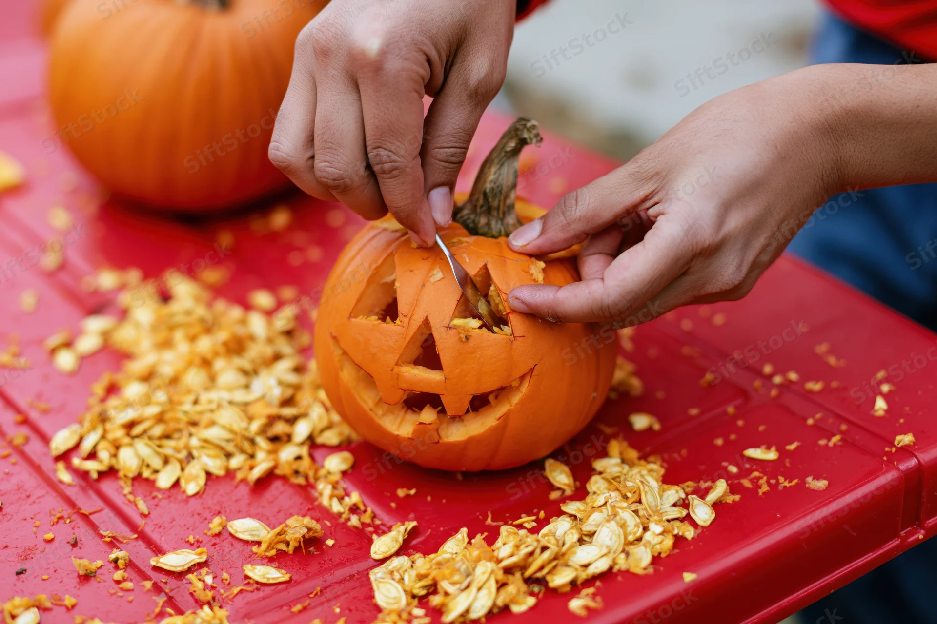 Hands carving a jack-o-lantern face into a small orange pumpkin on a red table surrounded by seeds