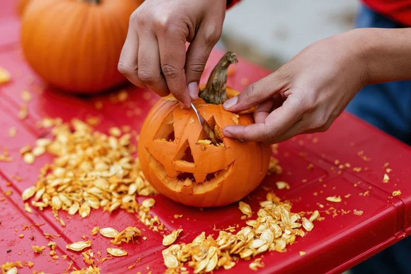 Hands carving a jack-o-lantern face into a small orange pumpkin on a red table surrounded by seeds