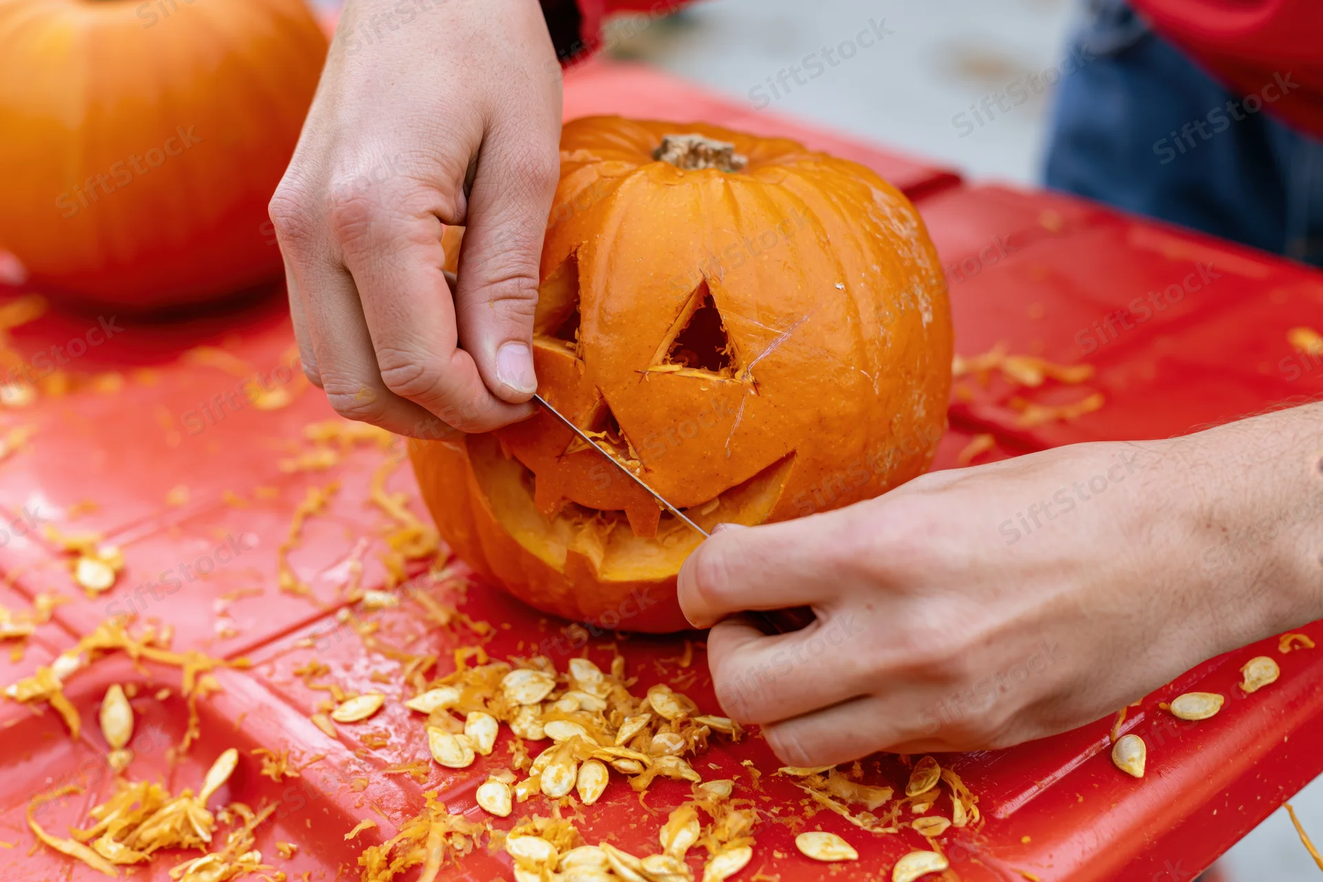 Hands carving a jack-o-lantern face into a small orange pumpkin on a red table