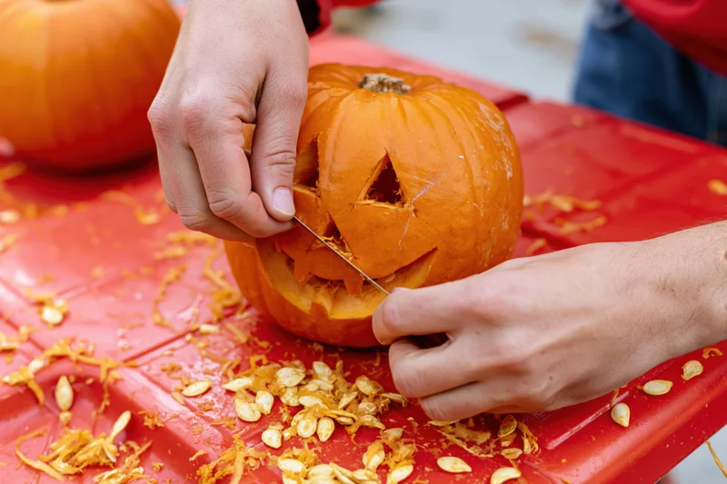 Hands carving a jack-o-lantern face into a small orange pumpkin on a red table