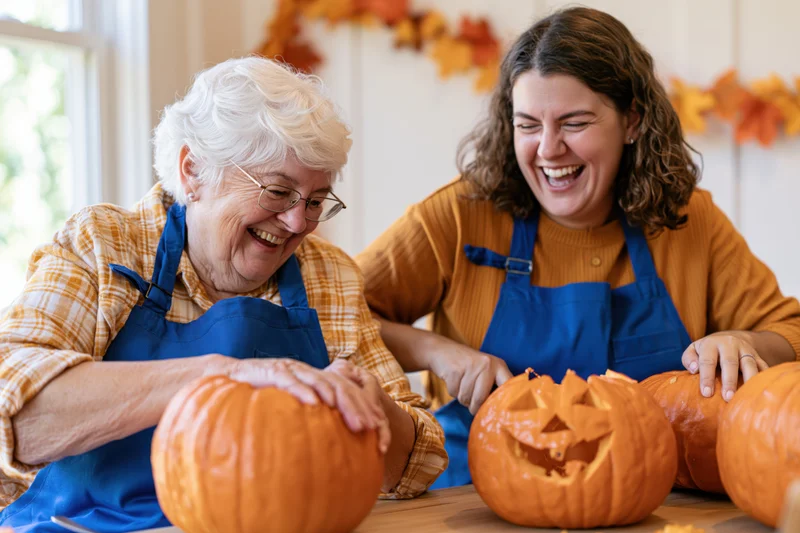 Senior woman and young woman laughing while carving pumpkins together at a kitchen table