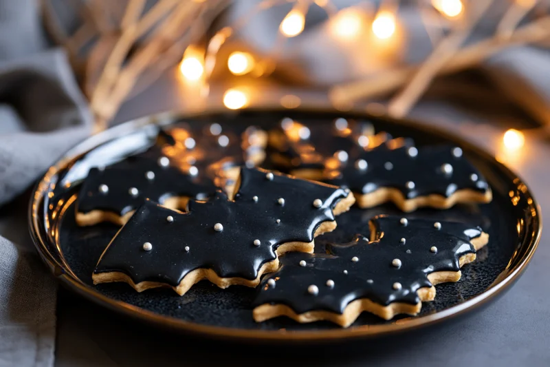Bat-shaped sugar cookies with black royal icing on a golden-rimmed plate, surrounded by warm string lights