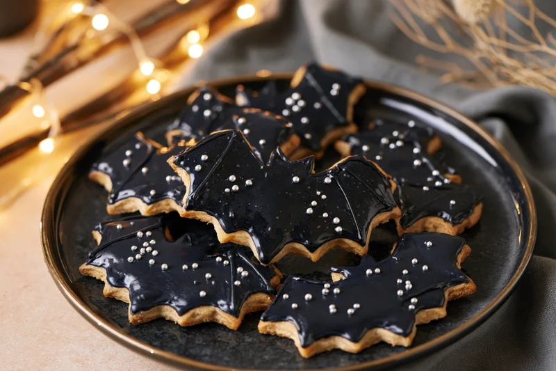 Bat-shaped sugar cookies with black royal icing on a dark plate surrounded by warm string lights