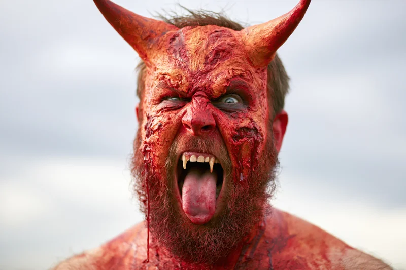 Bearded man in red devil costume makeup screaming with horns and fangs outdoors