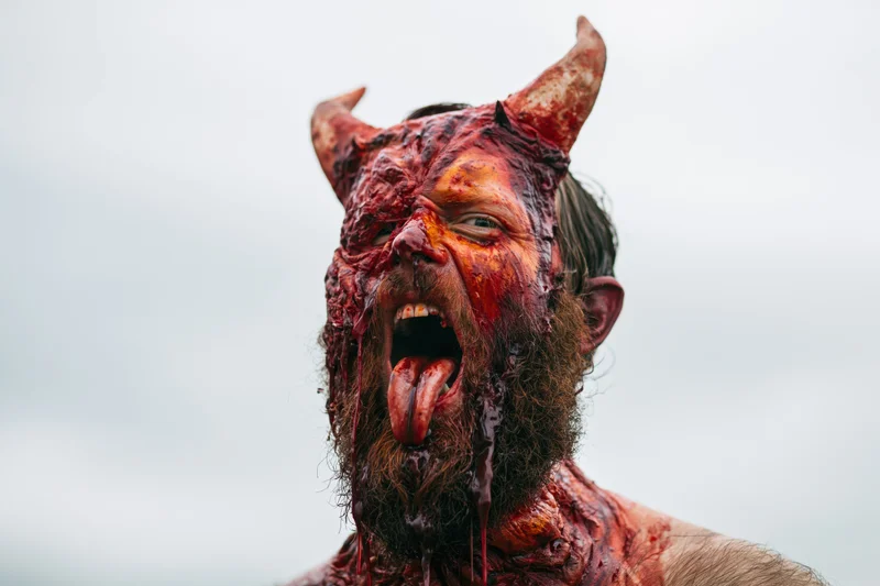 Bearded man in devil costume with bloodied makeup screaming outdoors