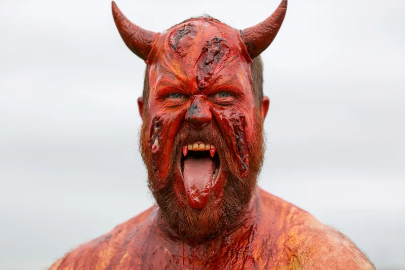 Bearded man in red devil costume makeup shouting with horns and fangs outdoors