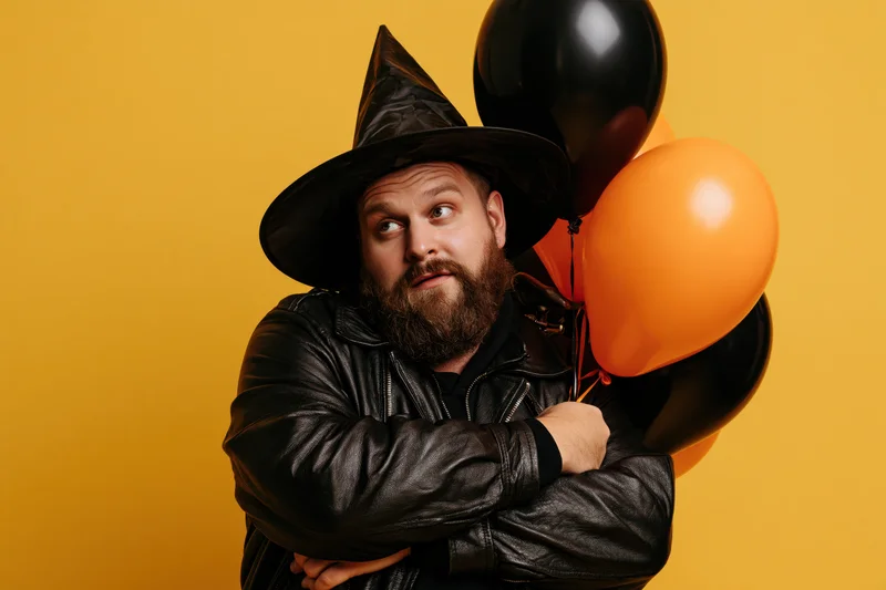 Bearded man in witch hat and leather jacket holding balloons, arms crossed, against yellow background