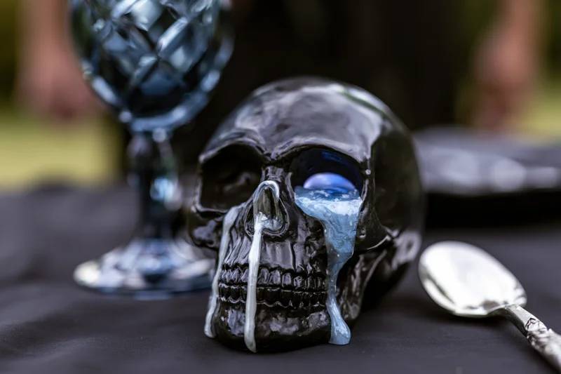 Black ceramic skull with melted wax dripping from eye and nose on dark table with glass goblet and spoon nearby