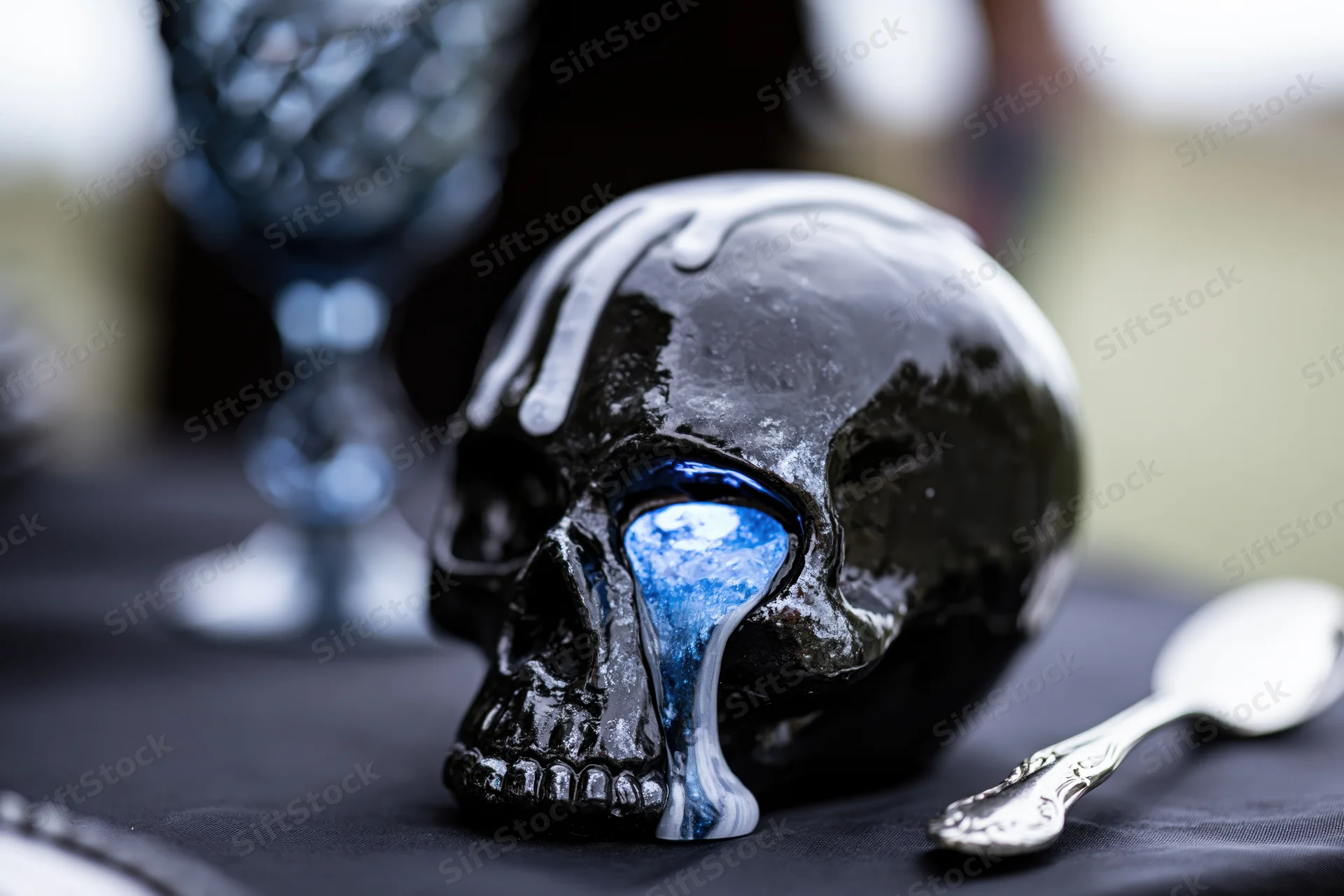 Black ceramic skull with melted white wax dripping on a dark table beside a spoon and glass goblet