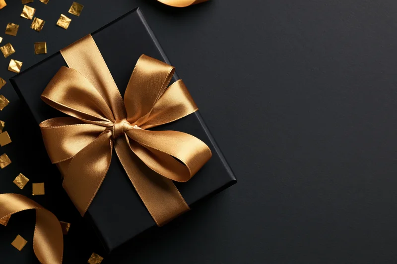 Black gift box with gold satin ribbon bow on dark surface, surrounded by gold confetti, top view