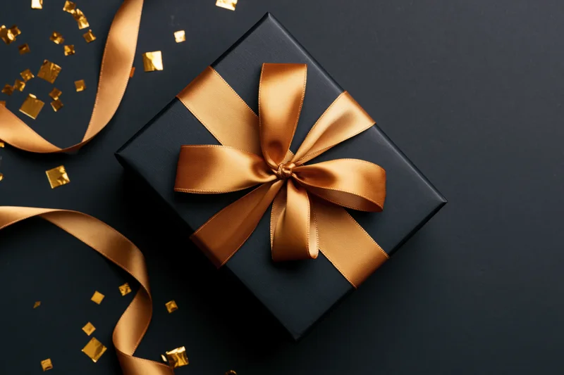 Black gift box with gold satin ribbon bow and scattered confetti on dark surface
