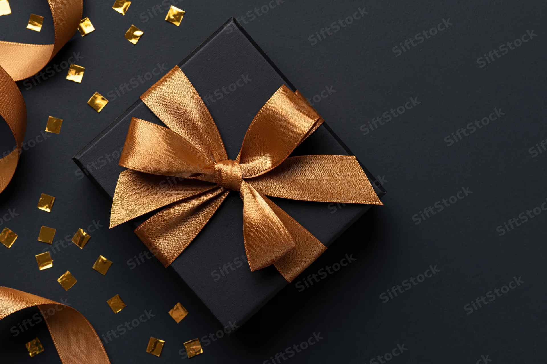 Black gift box with gold satin ribbon bow on dark surface surrounded by confetti