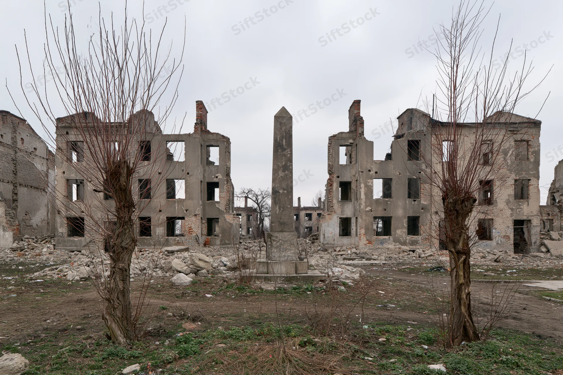 Bombed-out ruins of a city block with a central obelisk memorial surrounded by bare trees and rubble