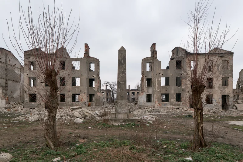Bombed-out ruins of a city block with a central obelisk memorial surrounded by bare trees and rubble