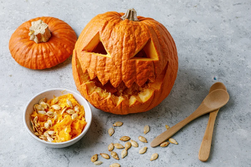 Carved jack-o-lantern pumpkin with triangle eyes and jagged mouth on gray countertop beside bowl of seeds and wooden spoons
